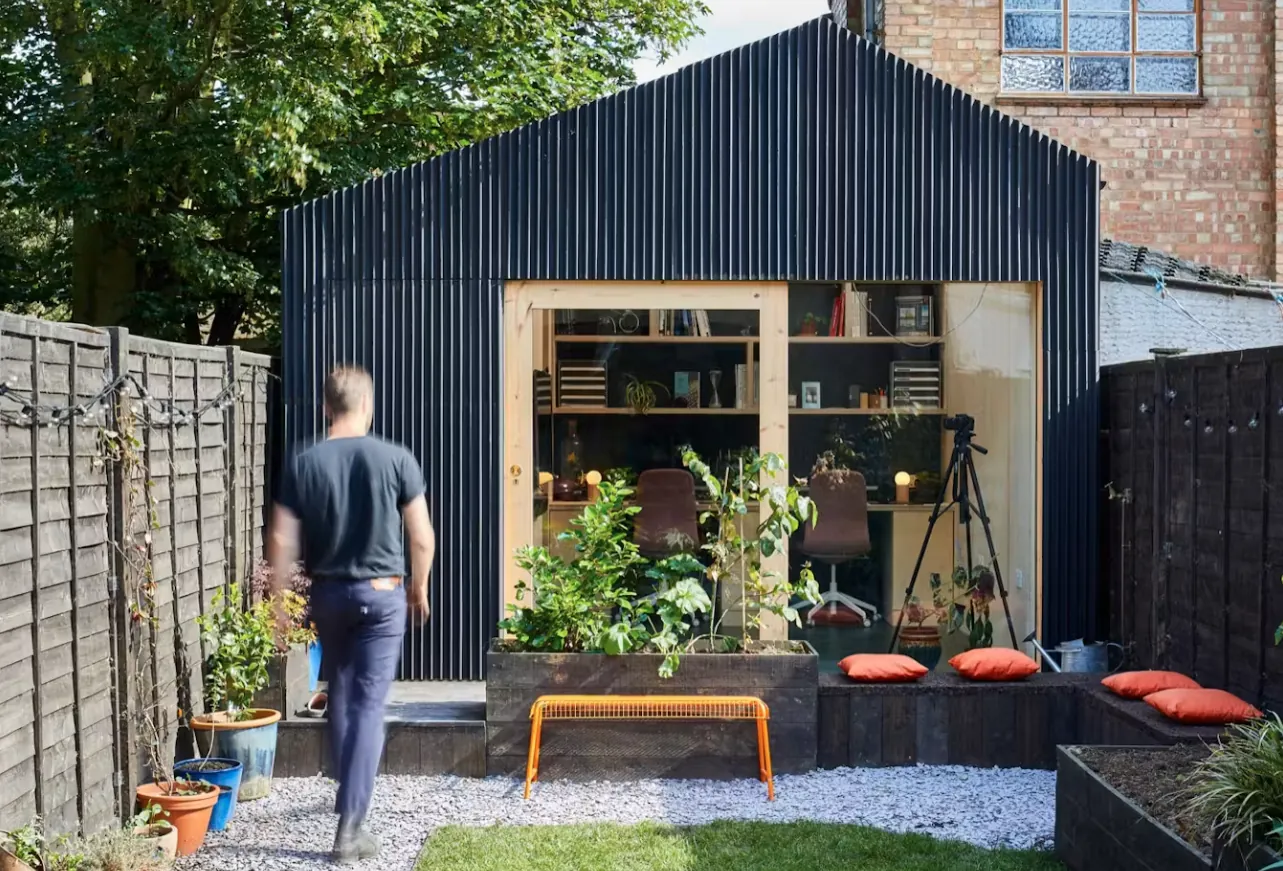 Dark corrugated backyard studio with its doors open onto a gravel garden and shelves inside