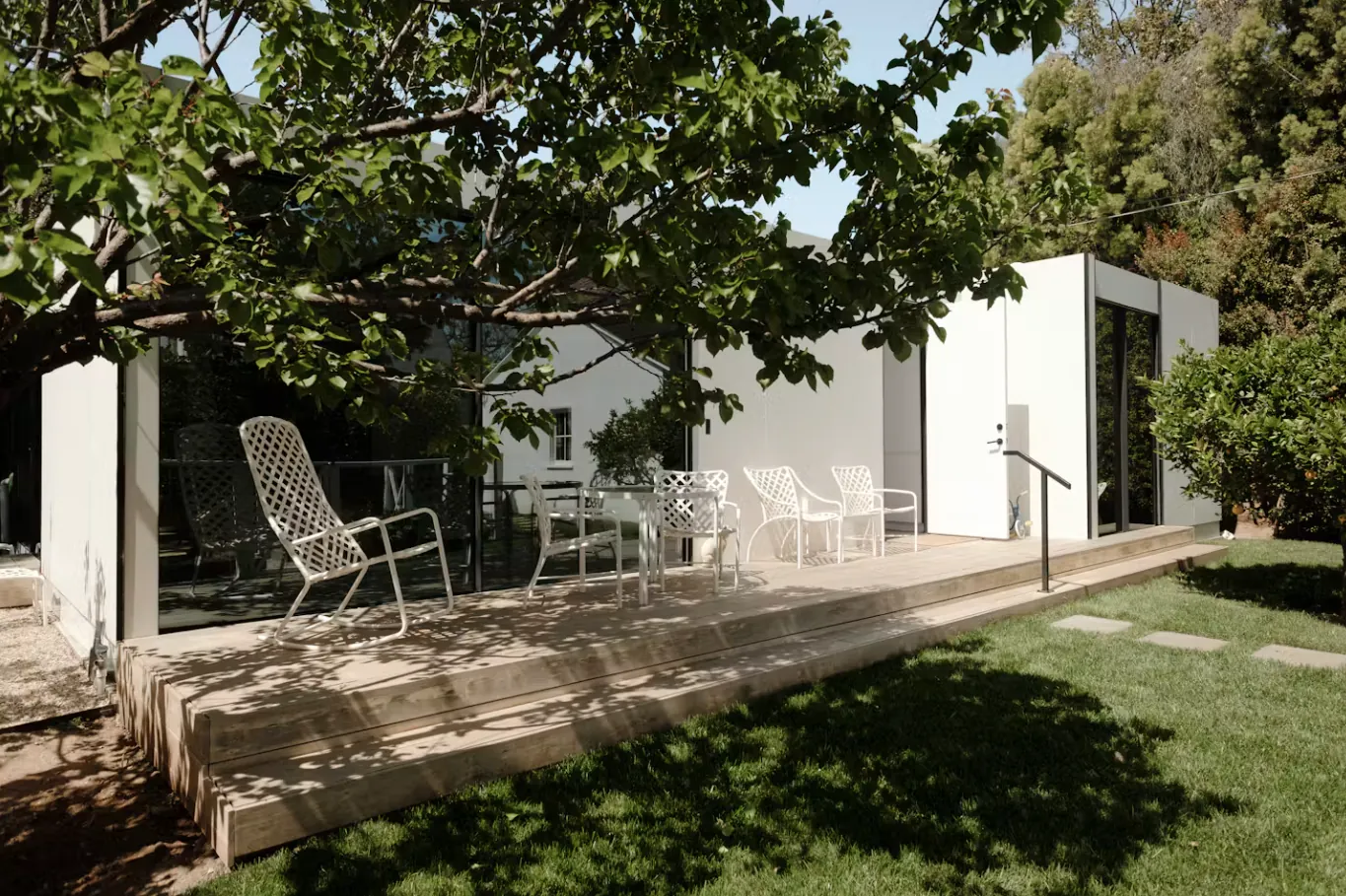 Single-storey white detached ADU with a wide deck and patio chairs under mature trees