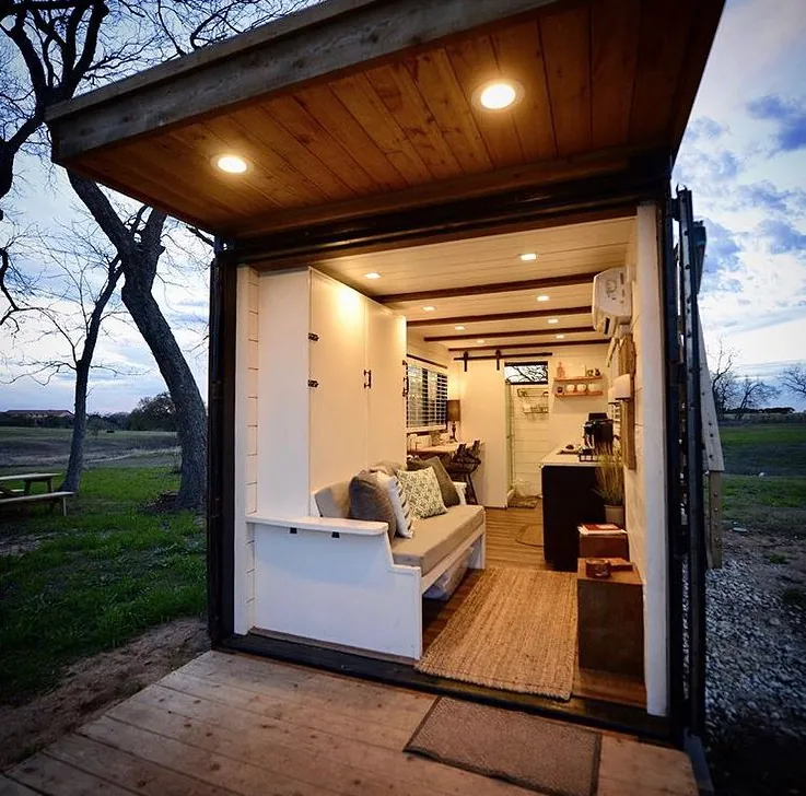 Tiny home with its glass end wall open, showing a bench seat, kitchen and wood ceiling inside